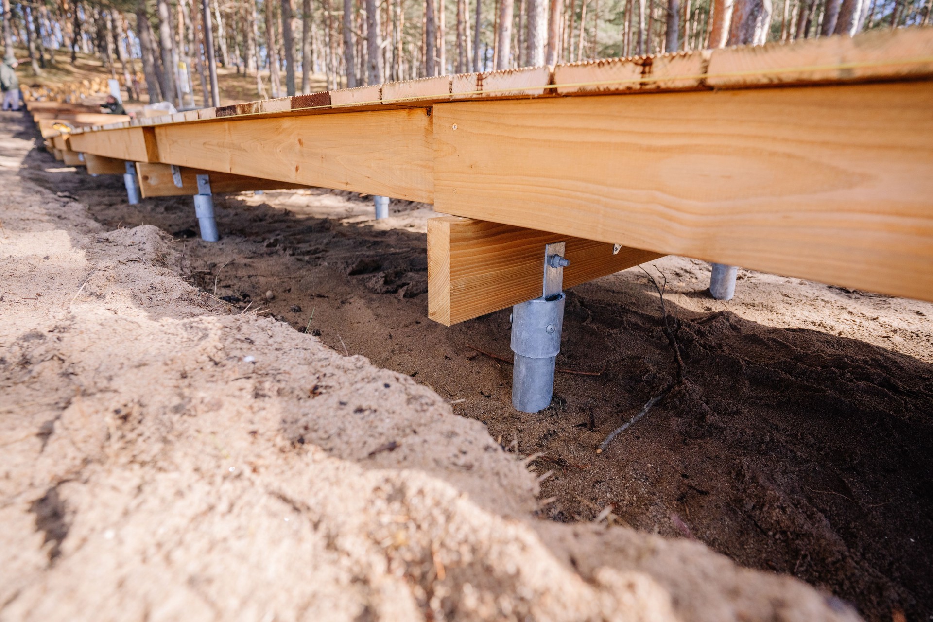 Wooden boardwalk structure supported by galvanized metal ground screws, built on sandy forest terrain, part of an outdoor construction project.