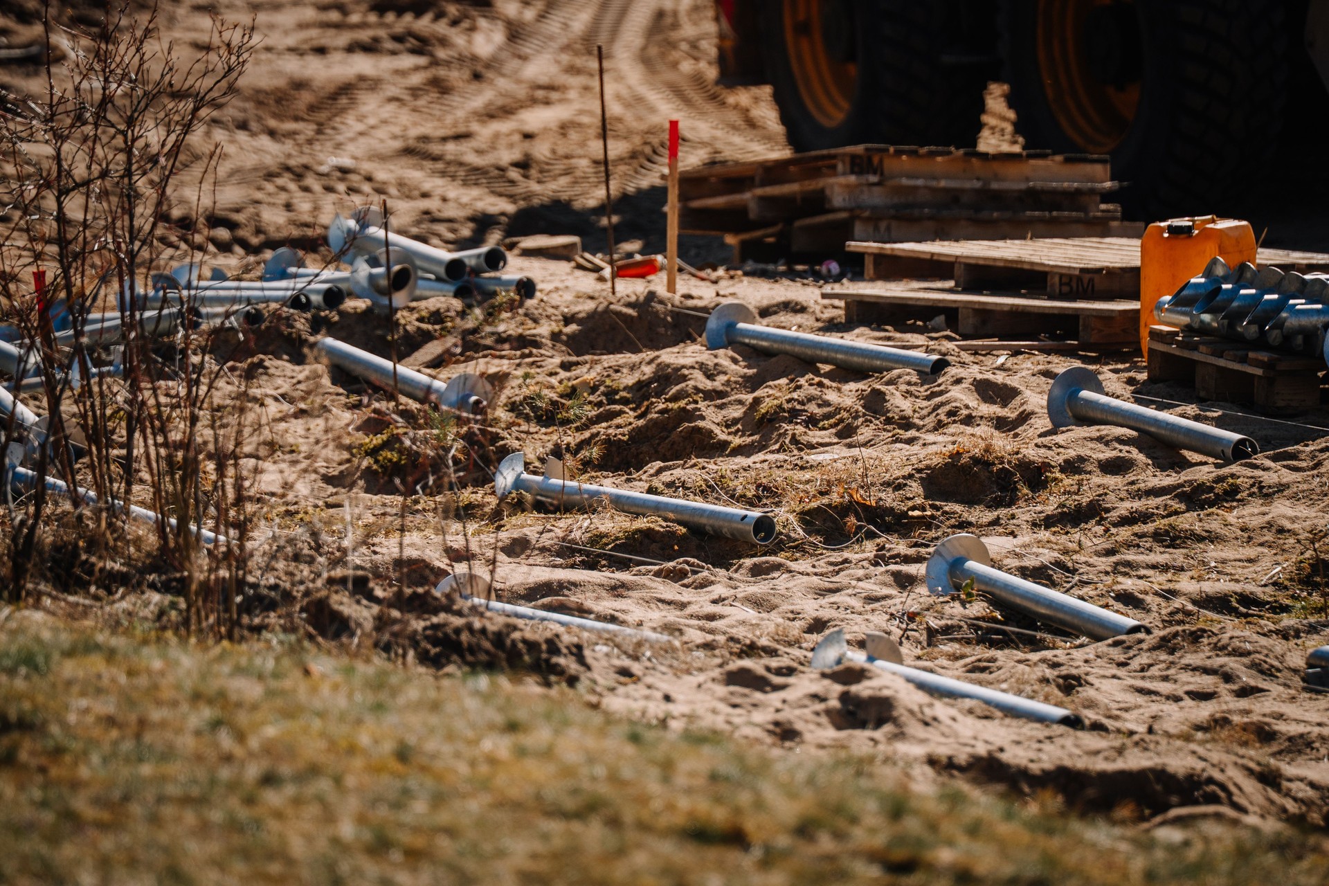 Scattered galvanized screw piles lie on sandy ground at a construction site, with machinery, pallets, and tools visible in the background.