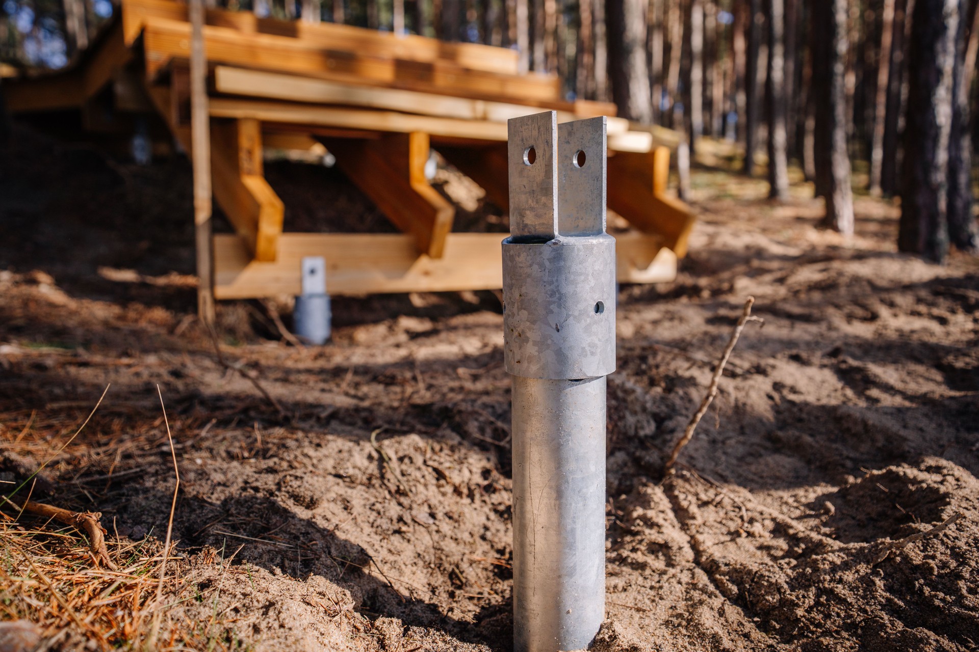 Close-up of a metal screw pile foundation anchored in sandy soil, with wooden stair construction in the blurred forest background on a sunny day.